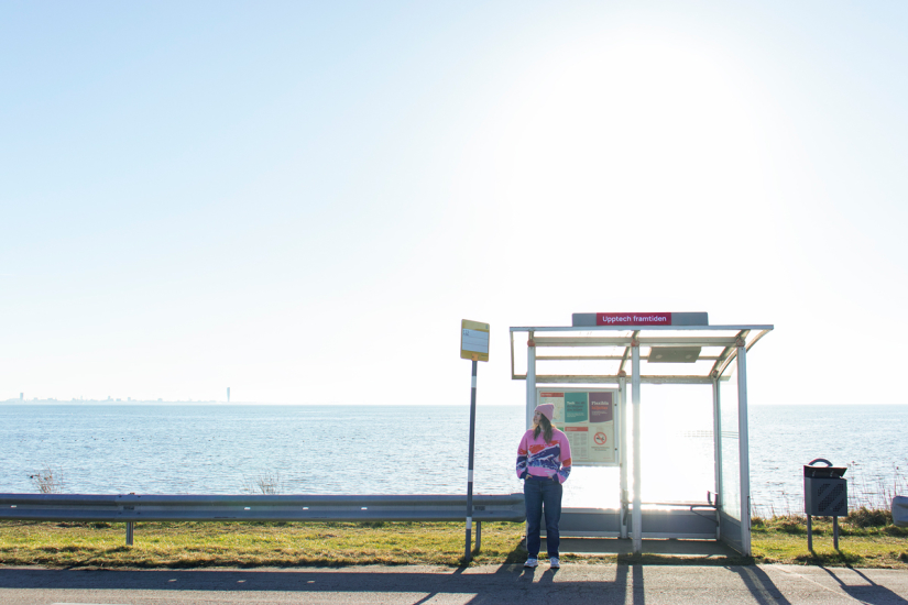 Foto på en kvinna som står vid en busshållsplats med solen i ryggen