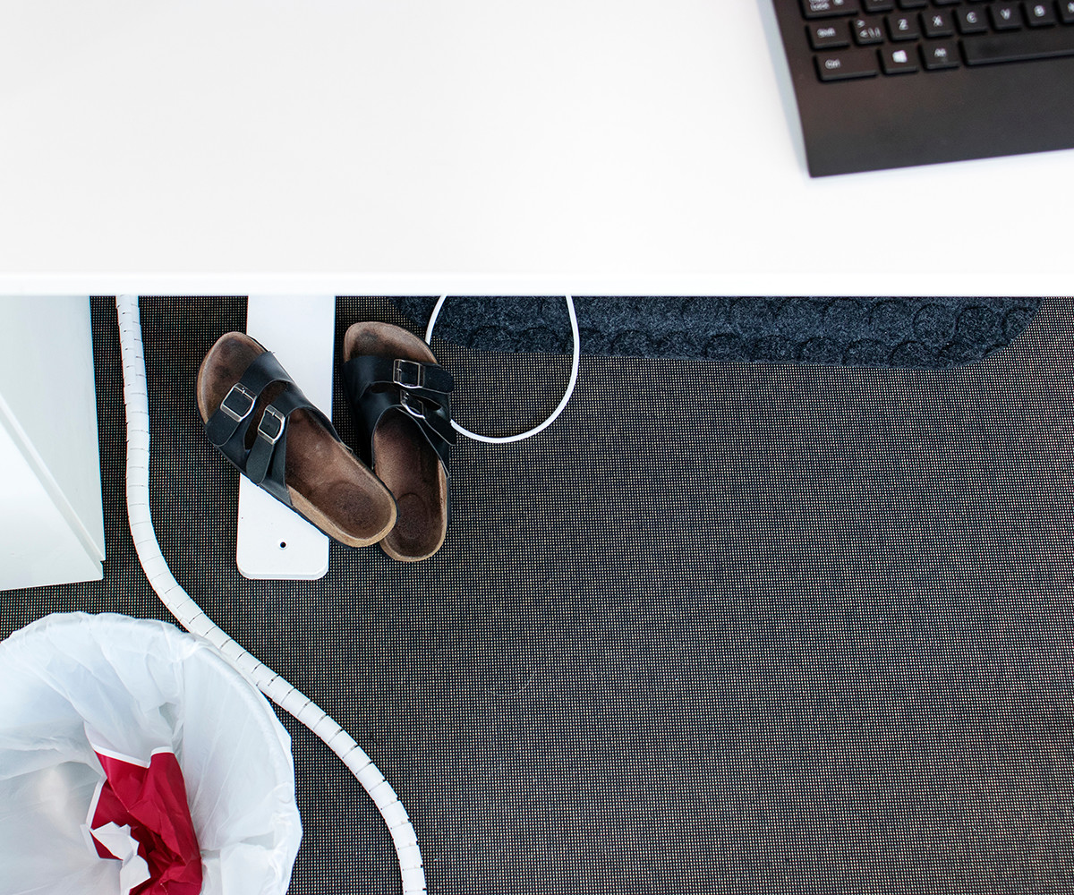 Foto på ett skrivbord med tofflor på golvet, sladdar och en soptunna intill.