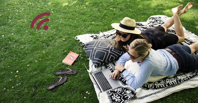 På bilden syns kvinnor som ligger på en filt på gräsmattan och framför sig har de en laptop som båda tittar på. Båda kvinnorna har solglasögon. En av dem har hatt.
