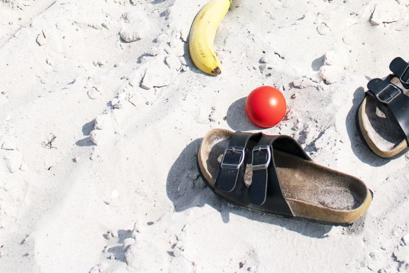 På bilden syns en sandstrand med två tofflor, en röd boll och en banan.
