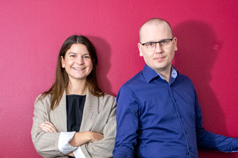 På bilden syns Tess Hamark och Karl Emil Nikka framför en röd vägg.