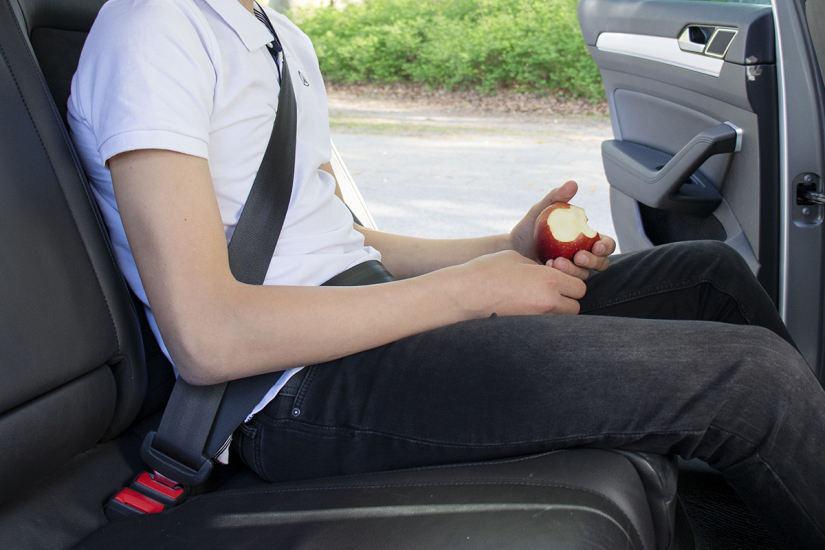 Bild på person som sitter i baksätet på en bil och känner sig säker och trygg med bilbälte. Personen håller ett äpple och har tagit några tuggor.