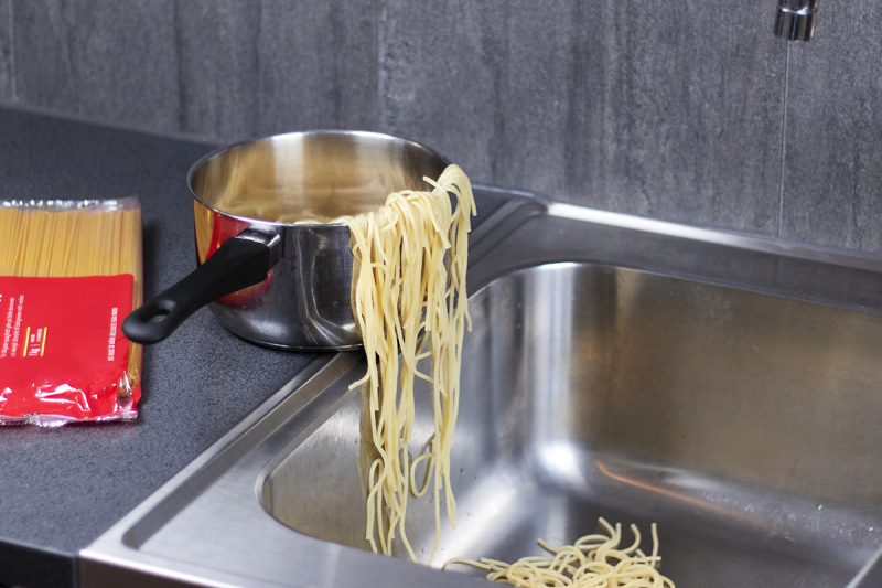 En kastrull med pasta vid vasken där en del av pastan åkt ner i vasken och resten hänger över kanten på kastrullen. Vid sidan om ligger ett paket okokt pasta.