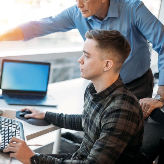 An image of an older man in a blue shirt directing a younger man in a checked shirt on how to run a programme on a desktop computer.