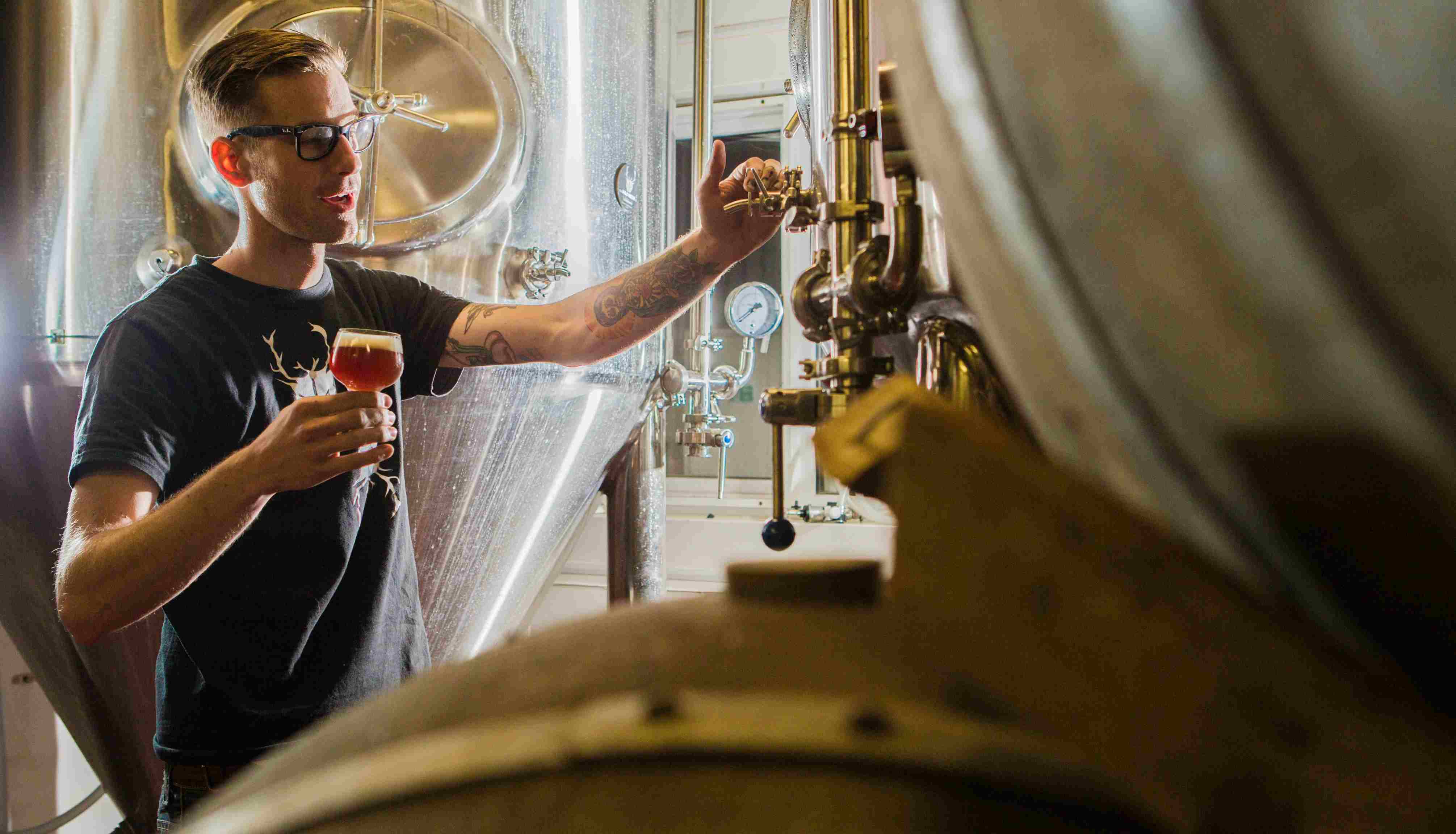 Man with glasses holding glass of beer adjusting equipment in a brewery