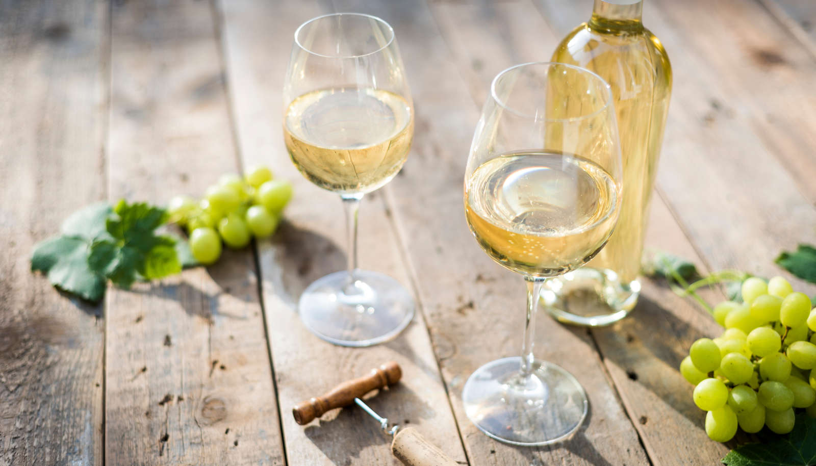 Two glasses of white wine in glasses, a bottle of white wine, white grapes and a corkscrew on a wooden table in the sunshine - natural wine 