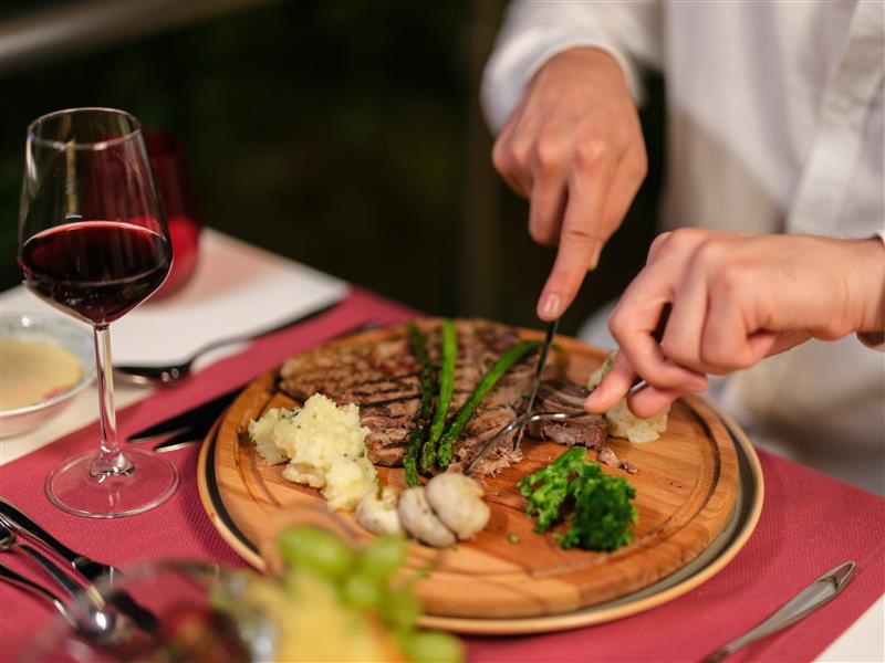 An image of a man cutting a steak meal, with mash potato and broccoli, and a glass of red wine on a table
