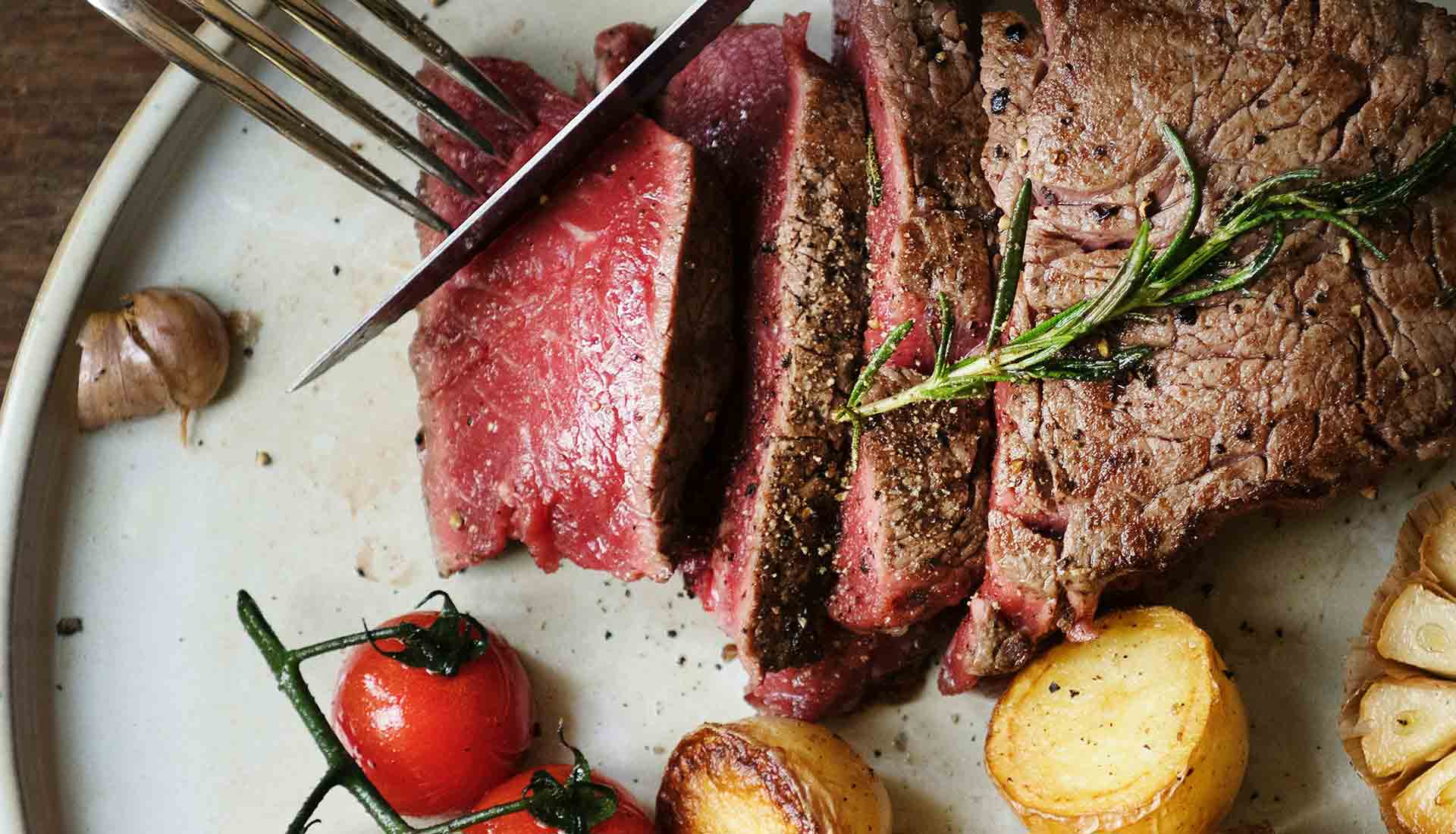 An image of a fillet of steak being cut, with roast potatoes and cherry tomatoes on a vine