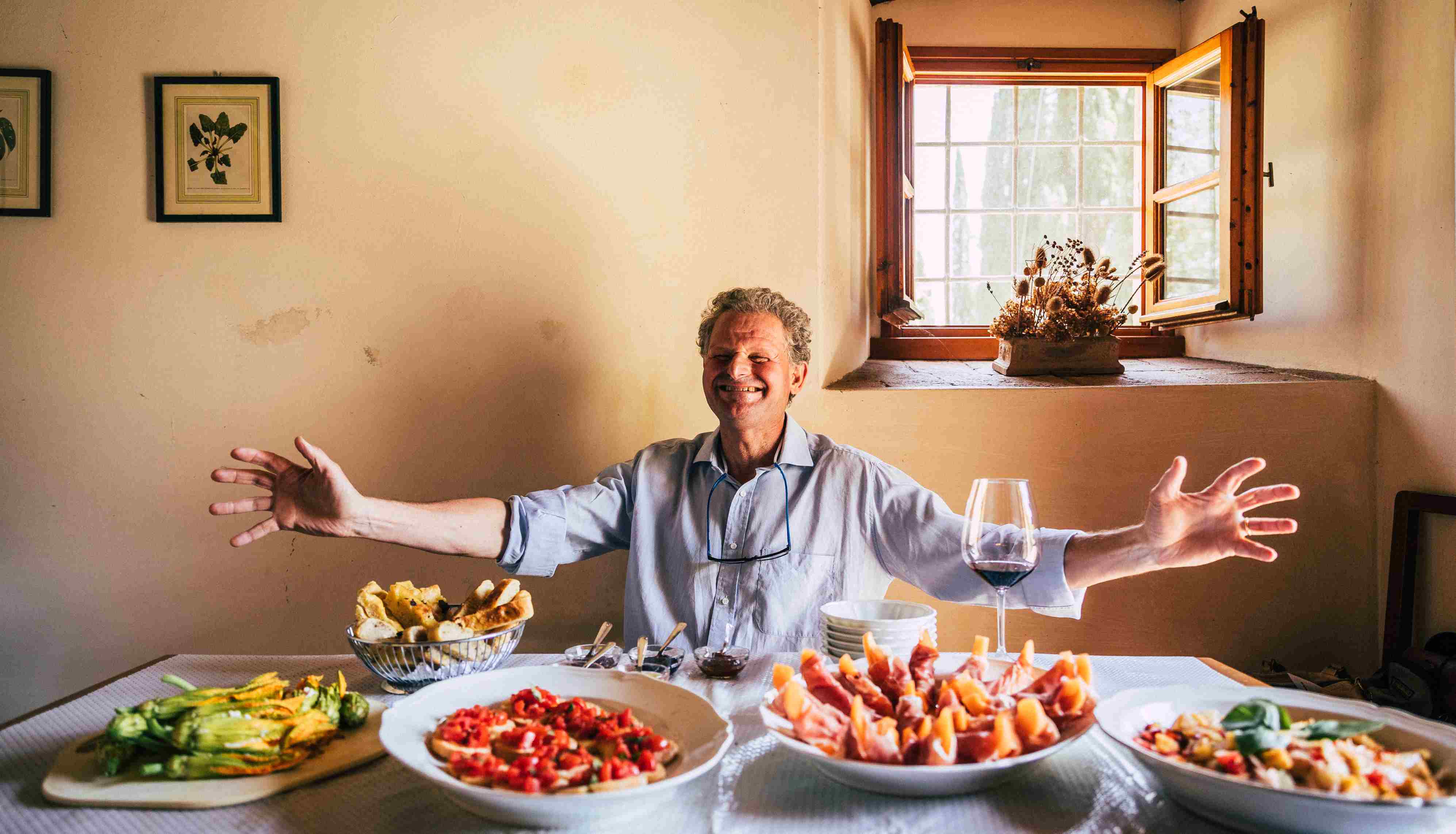 Italian man behind a table with wine and food welcoming with open arms