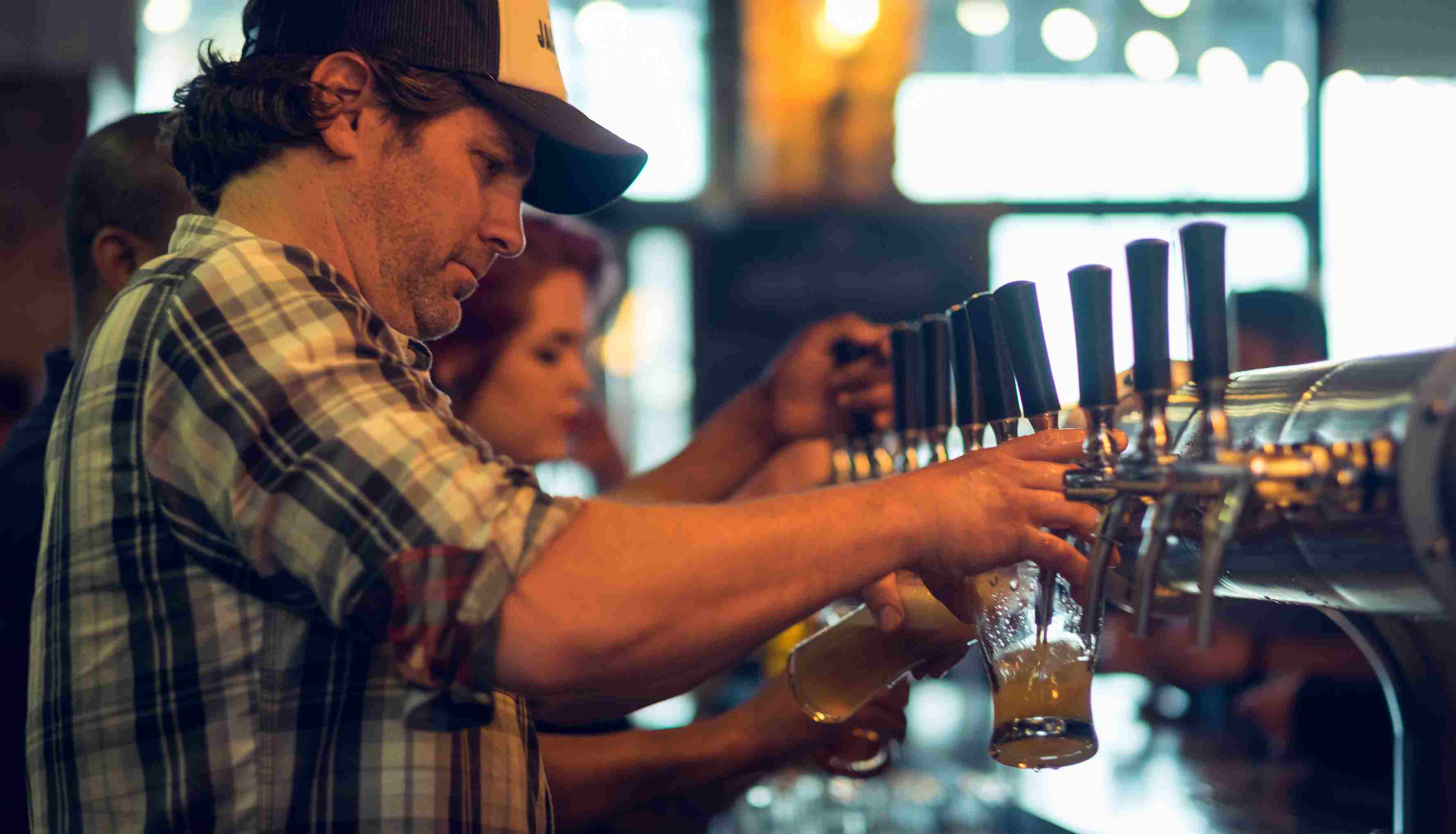 Bar workers bind a row of beer taps pouring beer, with a man with a cap in the foreground 