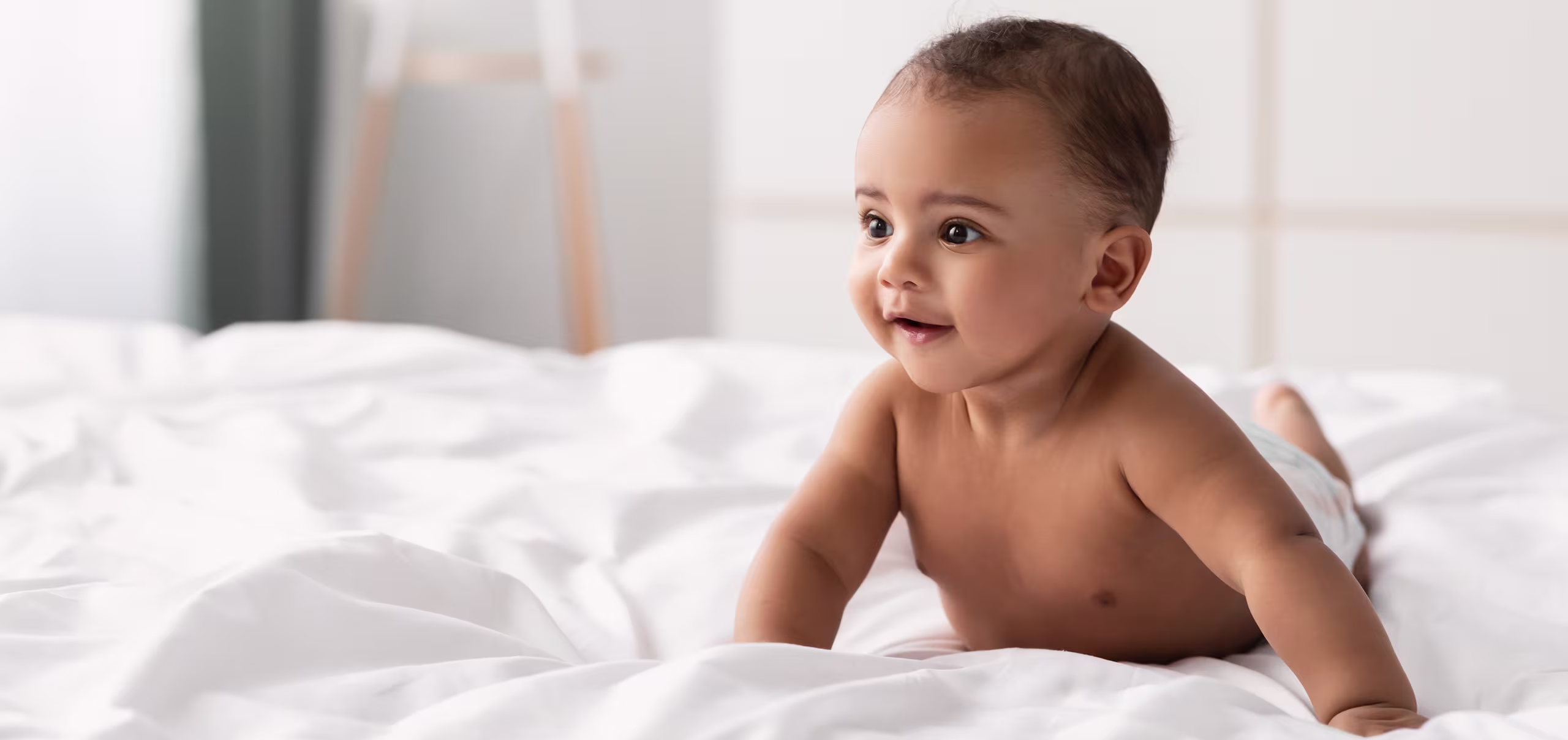 Guagua sonriente apoyado boca abajo sobre una cama con sábanas blancas.