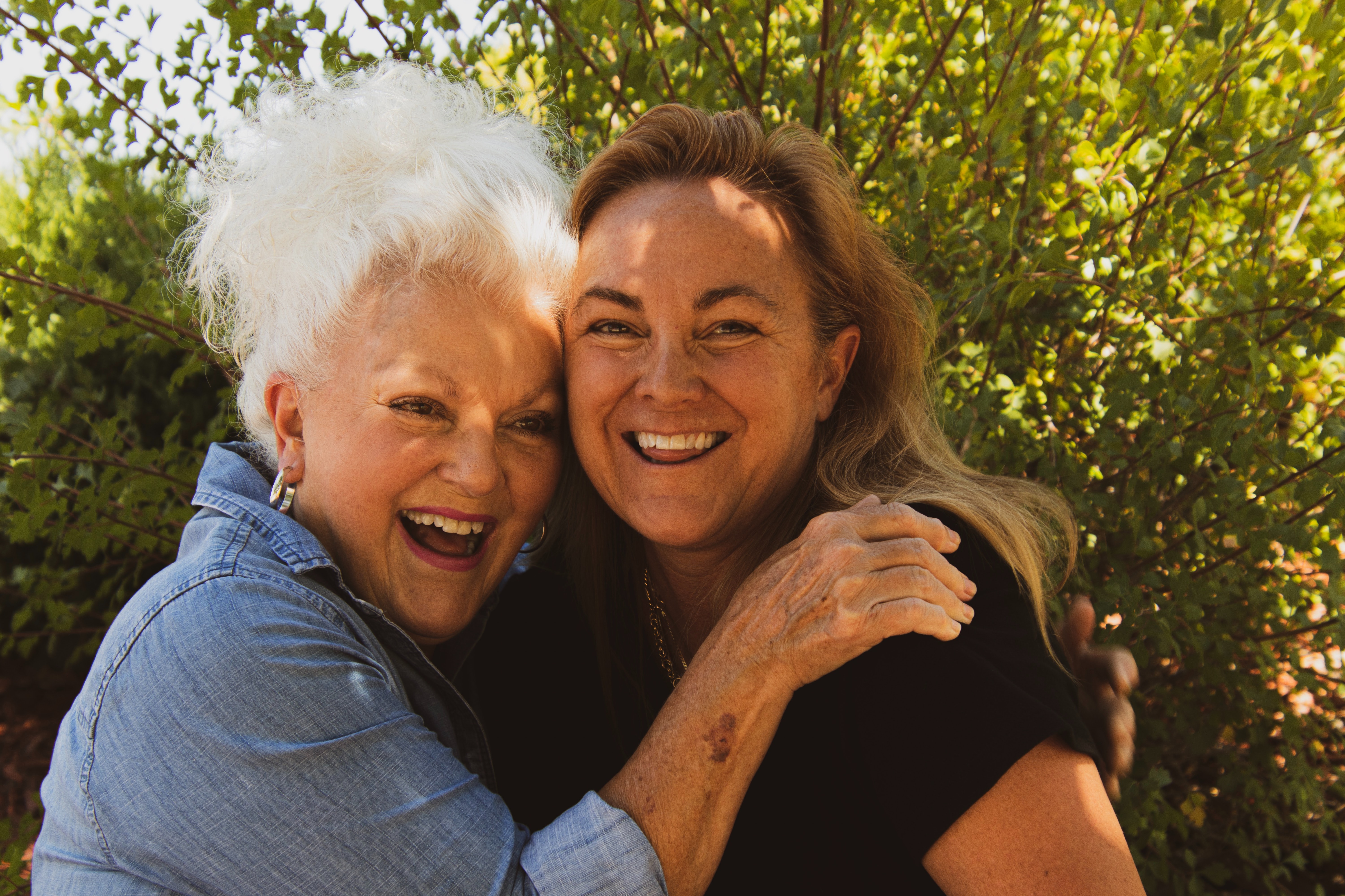 An older  white-haired woman wearing a blue button-up smiling and hugging an older  smiling woman wearing a black t-shirt. 