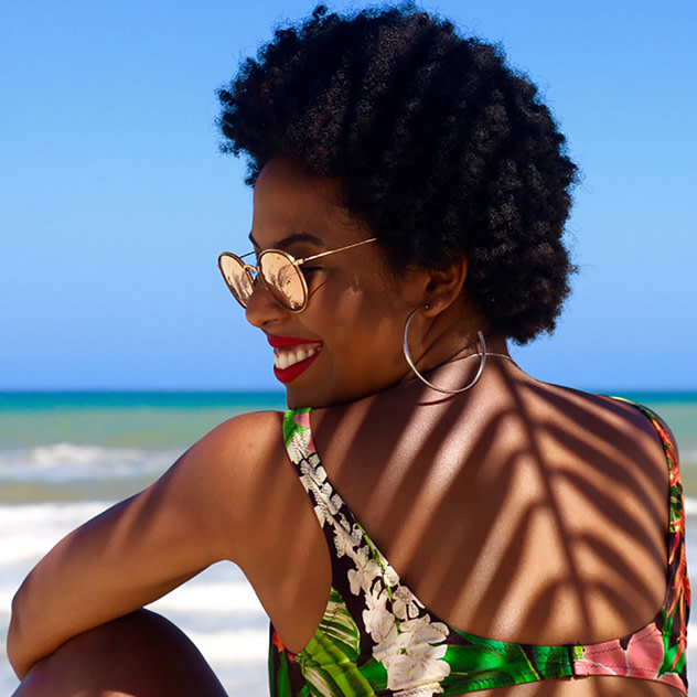 Eine Frau mit lockigem Haar und Sonnenbrille lächelt, während sie am Strand sitzt und das Meer im Hintergrund hat.