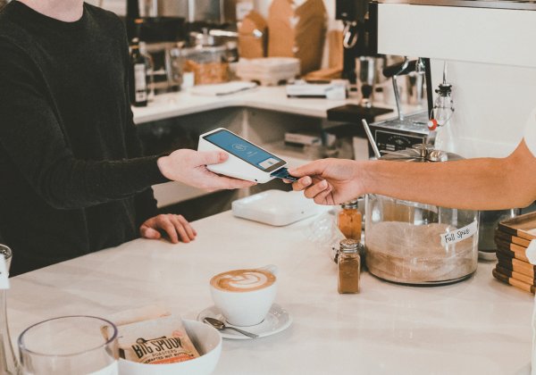 Es wird eine Bezahlsituation in einem Café gezeigt, in dem eine Person auf der rechten Seite die Bankkarte in ein kontaktloses Kartenlesegerät steckt, welches eine Person auf der linken Seite hält. Neben dem Kartenlesegerät steht ein Kaffee auf dem Tresen, im Hintergrund ist eine Kaffeemaschine und eine typische Kaffeeausstattung. 