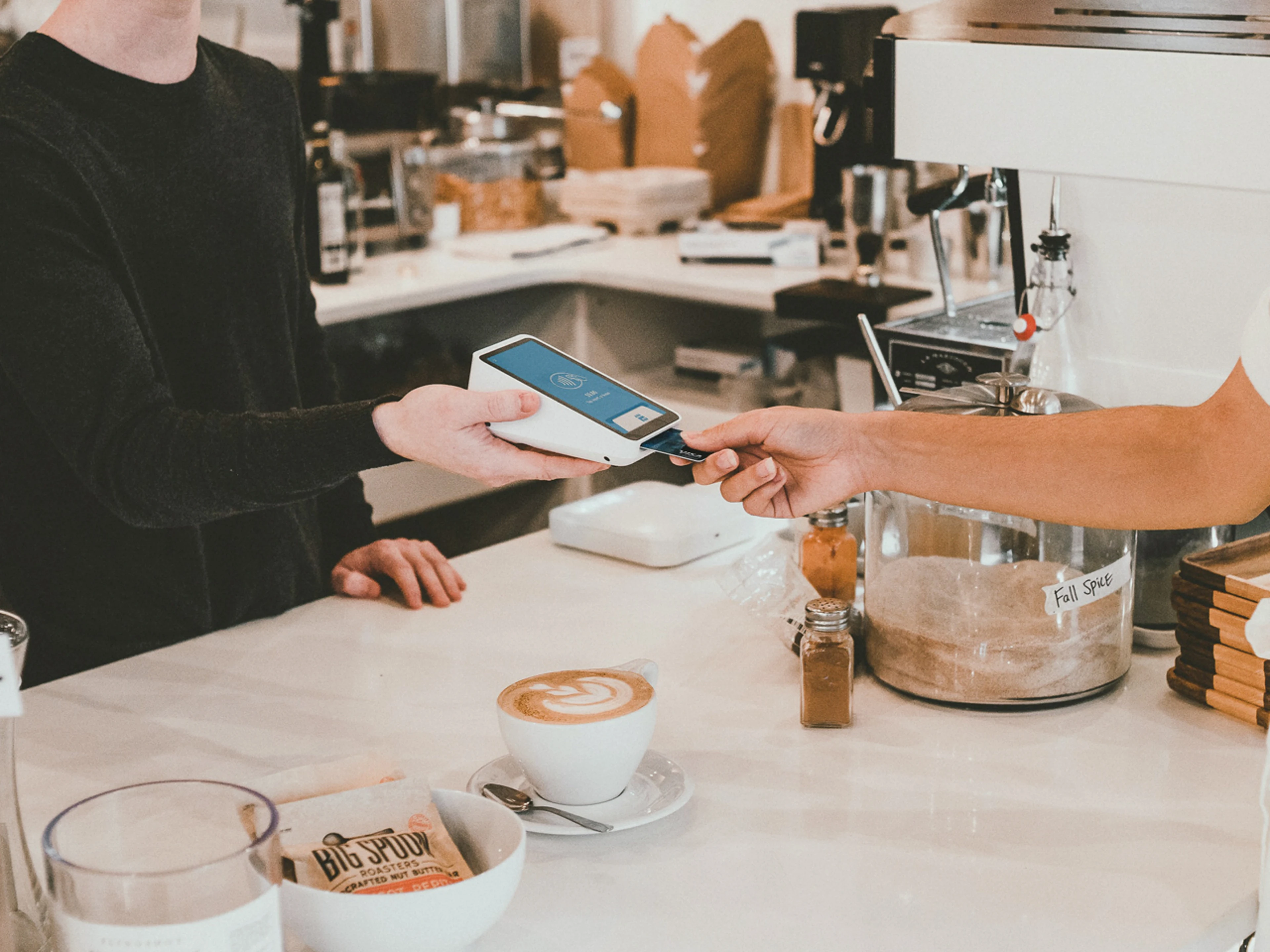 Es wird eine Bezahlsituation in einem Café gezeigt, in dem eine Person auf der rechten Seite die Bankkarte in ein kontaktloses Kartenlesegerät steckt, welches eine Person auf der linken Seite hält. Neben dem Kartenlesegerät steht ein Kaffee auf dem Tresen, im Hintergrund ist eine Kaffeemaschine und eine typische Kaffeeausstattung.