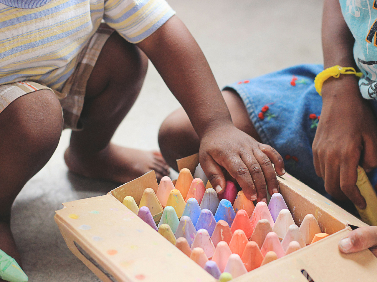 Ein Kind hockt und greift mit einer Hand in einen Karton mit bunter Kreide rein, der von einer anderen Person gehalten wird.