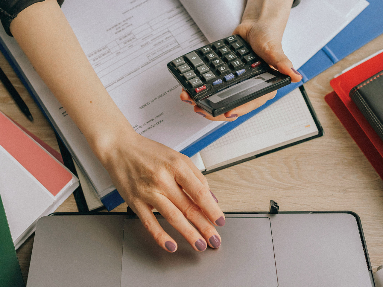 Zu sehen ist eine Hand, die auf dem Mousepad eines Laptos liegt und die andere Hand hält einen Taschenrechner. Und es liegen zusätzlich Hefter und Notizblöcke auf dem Schreibtisch.