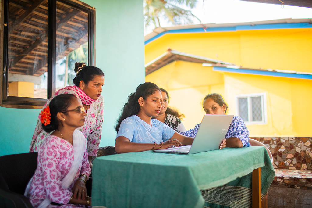 Five women of Indian origin are sitting on a terrace around a laptop. The woman in the center is typing something on the keyboard, while the others watch with interest. They are wearing colorful traditional clothing, and colorful houses bathed in sunlight can be seen in the background.