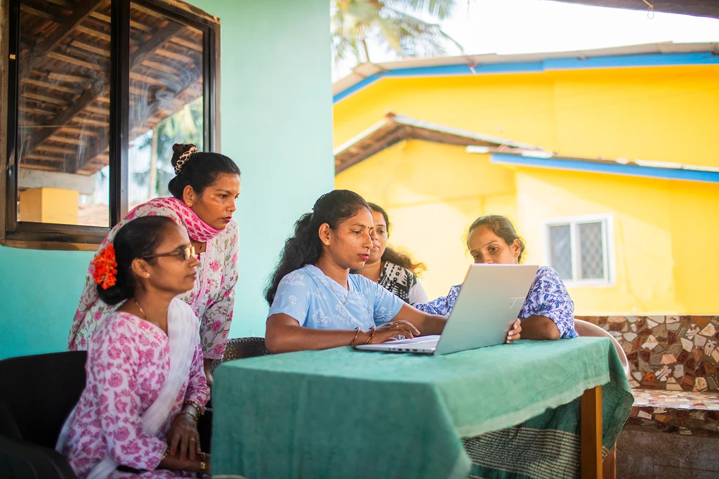 5 Frauen indischer Herkunft sitzen in auf einer Terrasse um ein Laptop. Die Frau im Zentrum tippt etwas auf der Tastatur, die anderen schauen interessiert zu. Sie tragen bunte, traditionelle Kleidung und im Hintergrund sind bunte, von der Sonne angestrahlte Wohnhäuser zu sehen.