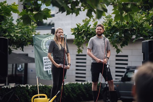 A woman and a man are standing on a stage. In front of them is a microphone stand. The woman is giving a speech. A Fridays for Future flag can be seen in the background. In front of them is a yellow watering can.