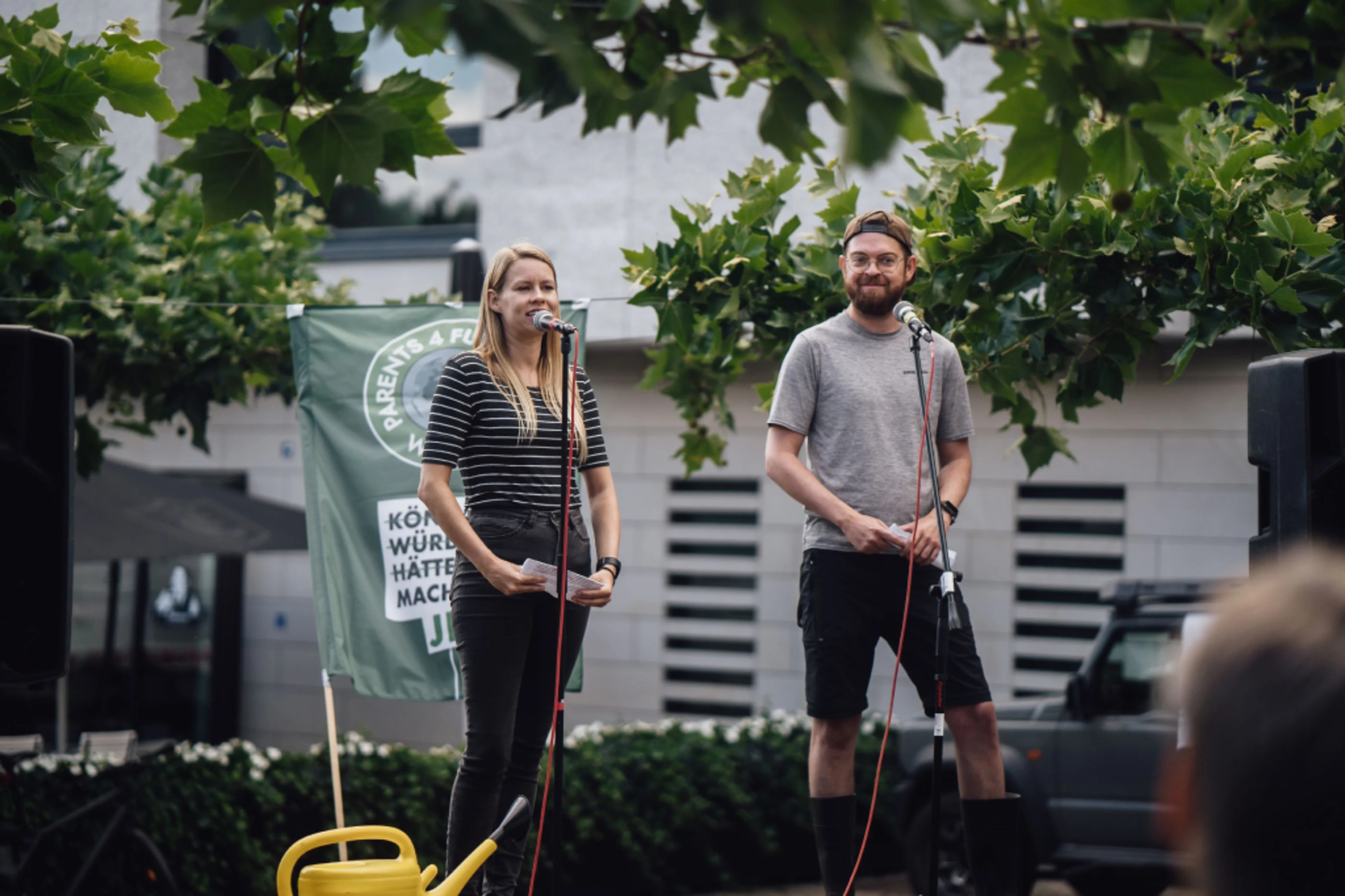 A woman and a man are standing on a stage. In front of them is a microphone stand. The woman is giving a speech. A Fridays for Future flag can be seen in the background. In front of them is a yellow watering can.