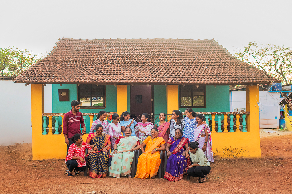 Fourteen women and one man have gathered in front of a small, colorfully painted house for a group photo. Almost all of the women are wearing traditional Indian saris. They are laughing cheerfully at the camera or looking at each other. 