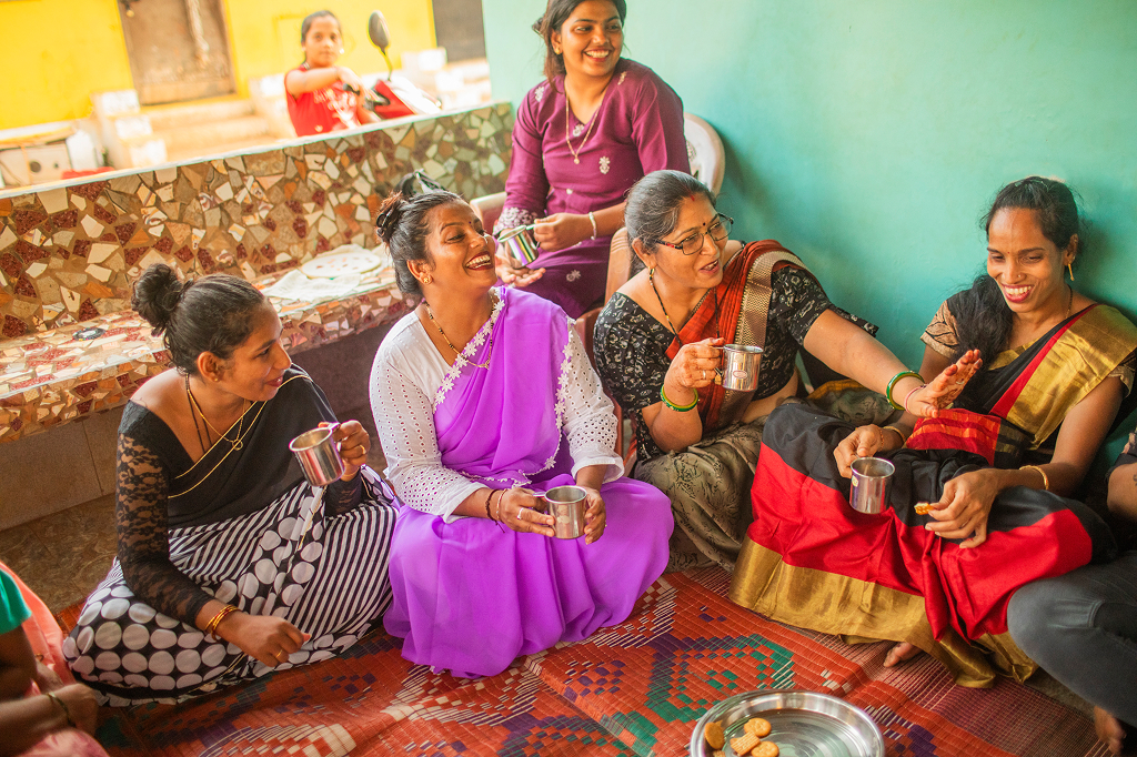 Five women of Indian origin sit cross-legged in a colorful living room with a red carpet and turquoise walls. They drink chai from silver cups and chat, laughing and gesturing.