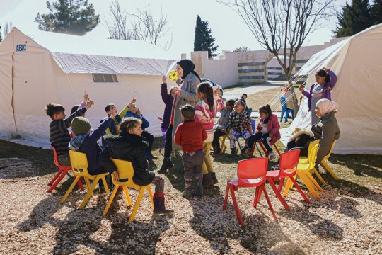Children in a circle of chairs, around a teacher. In the background are tents. 