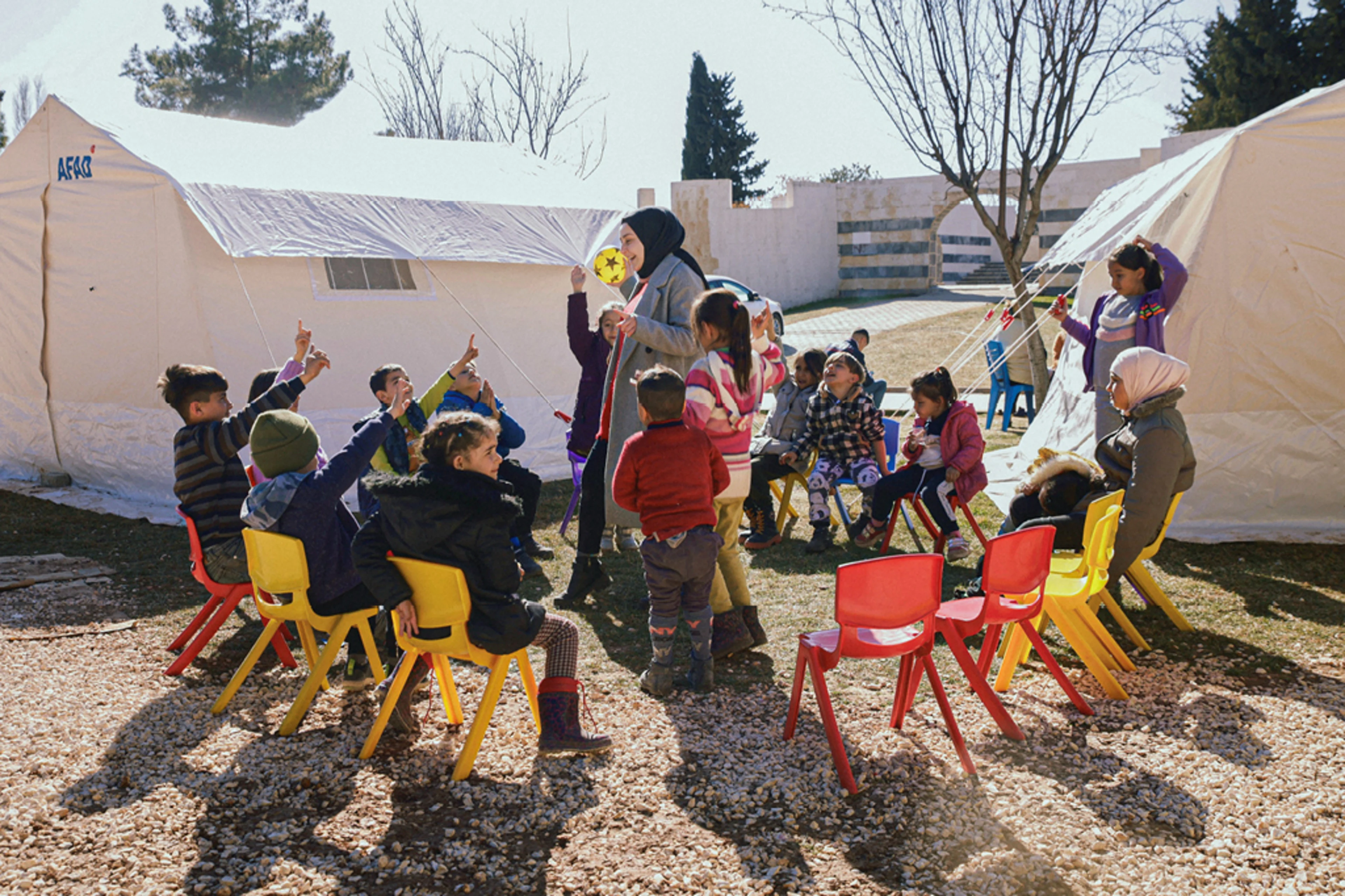 Children in a circle of chairs, around a teacher. In the background are tents.