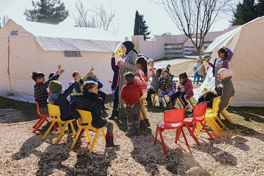 Kinder in einem Stuhlkreis, um eine Lehrerin. Im Hintergrund sind Zelte.