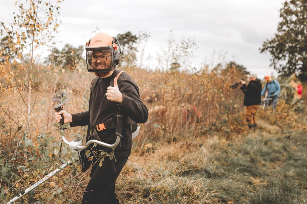 Ein Mann steht in einer herbstlichen Moorlandschaft. Er trägt einen Helm und Gehörschutz und trägt einen Kantschneider vor sich her, mit dem er die Vegetation trimmt. Er blickt lachend in die Kamera und zeigt mit einer Hand "Daumen hoch". Im Hintergrund befinden sich 3 andere Menschen, die teilweise Arbeiten verrichten und Fotos machen.