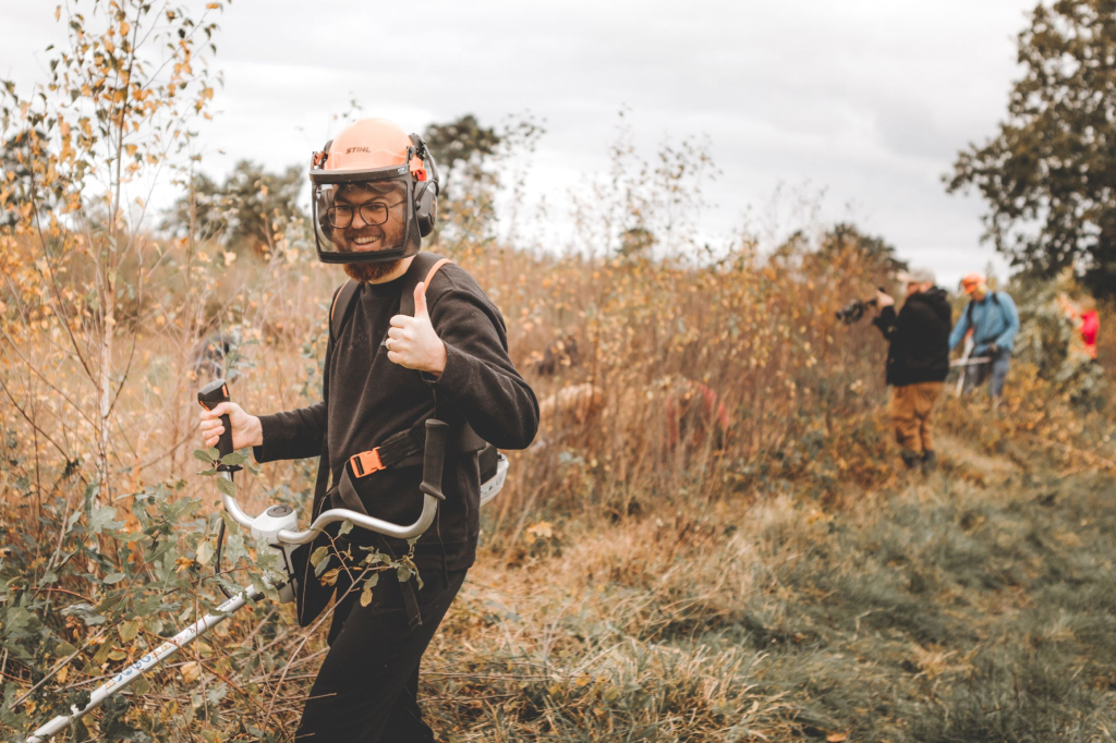 A man stands in an autumny moorland landscape. He wears a helmet and ear protection and carries a brush cutter in front of him, which he uses to trim the vegetation. He looks at the camera with a smile and gives a thumbs up with one hand. In the background, there are three other people, some of whom are working and taking photos.