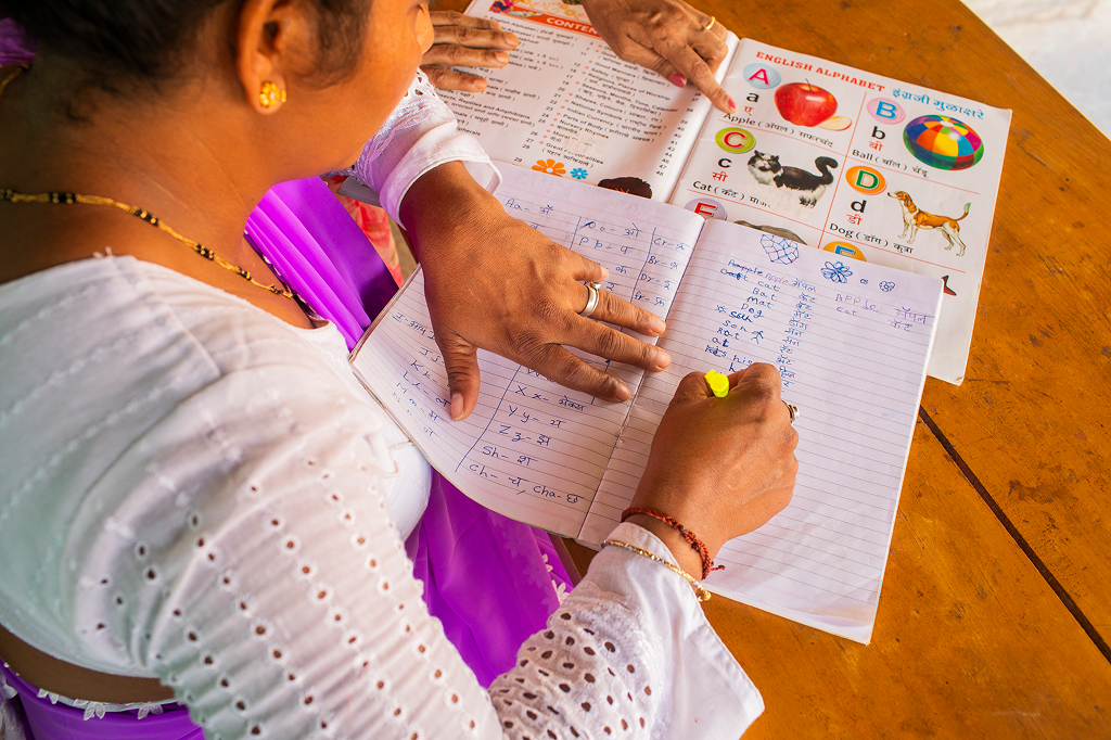 A woman of Indian origin writes vocabulary from an English-Hindi workbook in her notebook. On the left-hand page, she has written down the Latin alphabet, and on the right-hand page, she writes down simple words such as “apple,” “ball,” “cat,” and “dog.” We look over her shoulder; she is wearing a colorful traditional garment and lots of jewelry. Another pair of hands points to individual illustrations in the workbook.