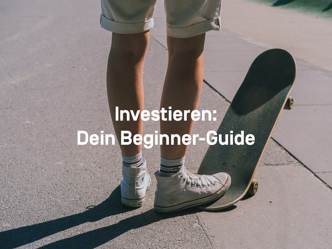 Ein junger Mann in kurzer Hose und Chucks steht auf der Straße. Sein rechter Fuß steht auf dem hinteren Teil eines Skateboards, die vorderen Rollen schweben in der Luft. 
Text: Investieren: Dein Beginner-Guide
