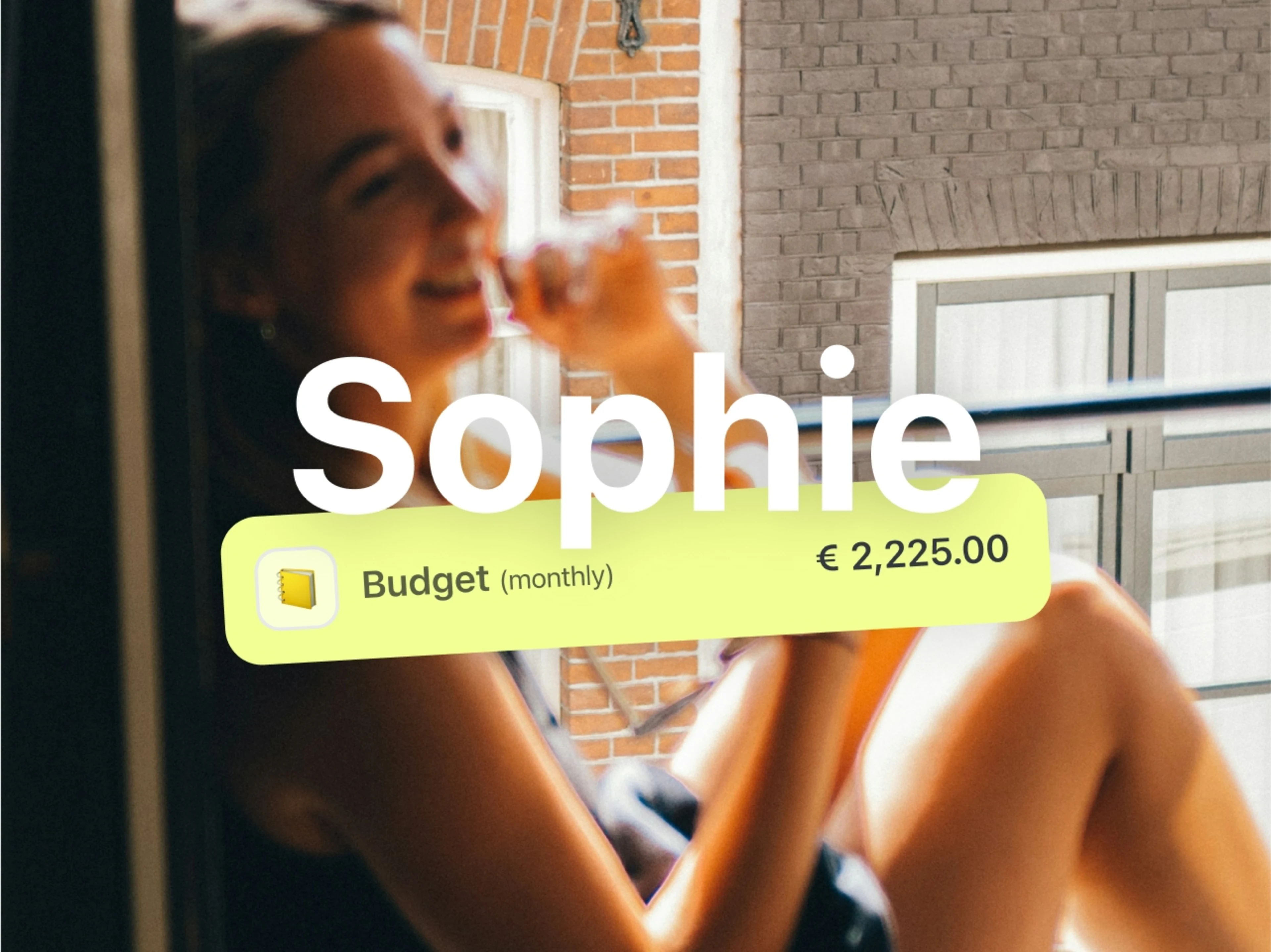 The name Sophie is written large in the center of the motif, with a block of text underneath:
Budget (monthly) €2,225.00. Behind it is a photo.
In the foreground, a young woman is sitting on a windowsill and laughing. The focus is on the wall of the house behind her, where we see old windows and a red brick wall.