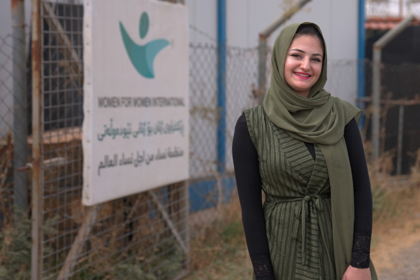 Zu sehen ist eine Teilnehmende des Programms im Irak, sie lächelt und steht vor dem Gebäude in dem die Schulungen stattfinden. 