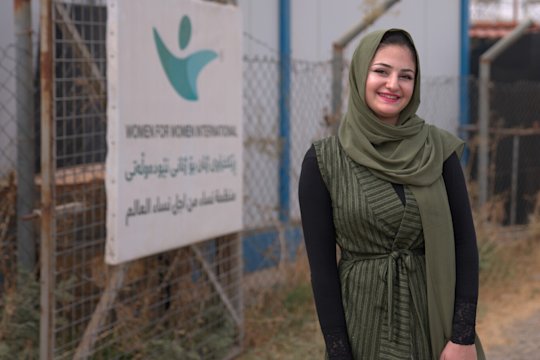 Zu sehen ist eine Teilnehmende des Programms im Irak, sie lächelt und steht vor dem Gebäude in dem die Schulungen stattfinden.