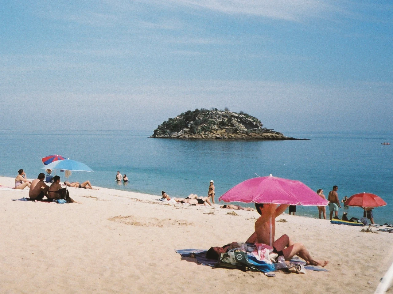 Zu sehen ist ein Sandstrand mit Blick auf das Meer und einer kleinen Insel im Meer. Ein paar Menschen sitzen mit Sonnenschirmen im Sand.