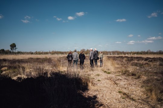 6 Personen stehen mit dem Rücken zum Betrachter gewandt in einer Landschaft. Die Landschaft ist karg und trocken. Der Himmel ist blau mit vereinzelten Wolken.