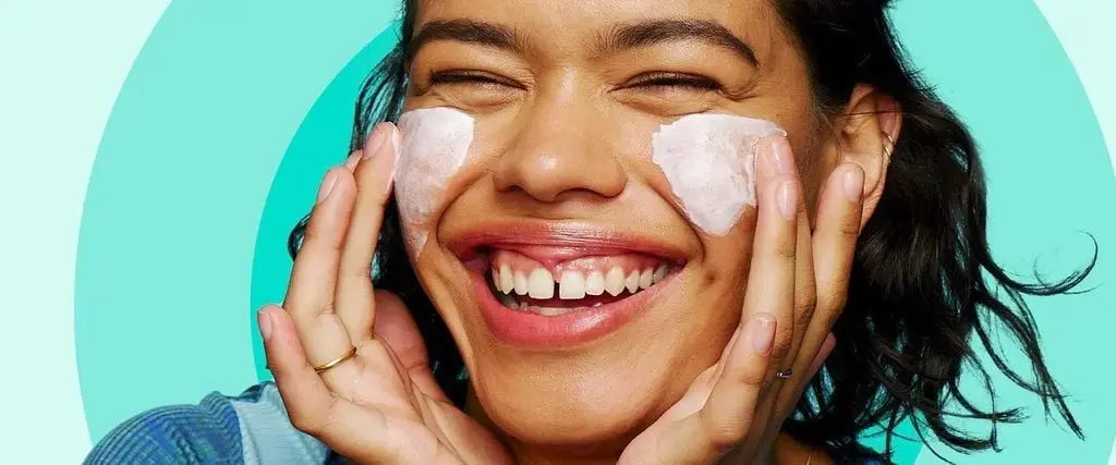 A smiling person applies cream to their cheeks with both hands, surrounded by a turquoise background, while wearing a blue patterned shirt.