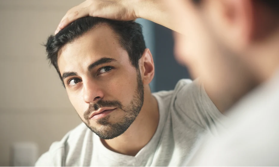 Man with dark brown hair and beard checking his hairline in the mirror.