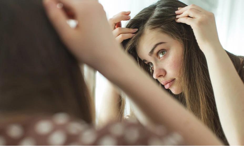 Woman with long, light brown hair checking her part in the mirror.