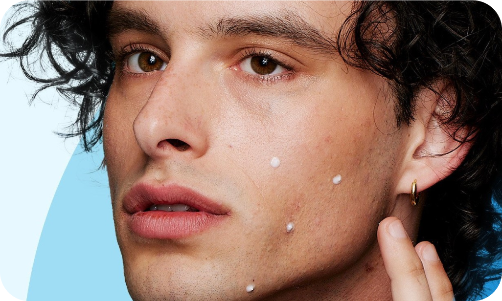 A person with curly hair applies small white dots of cream on their face, focusing intently. The background features light blue and darker blue curves.