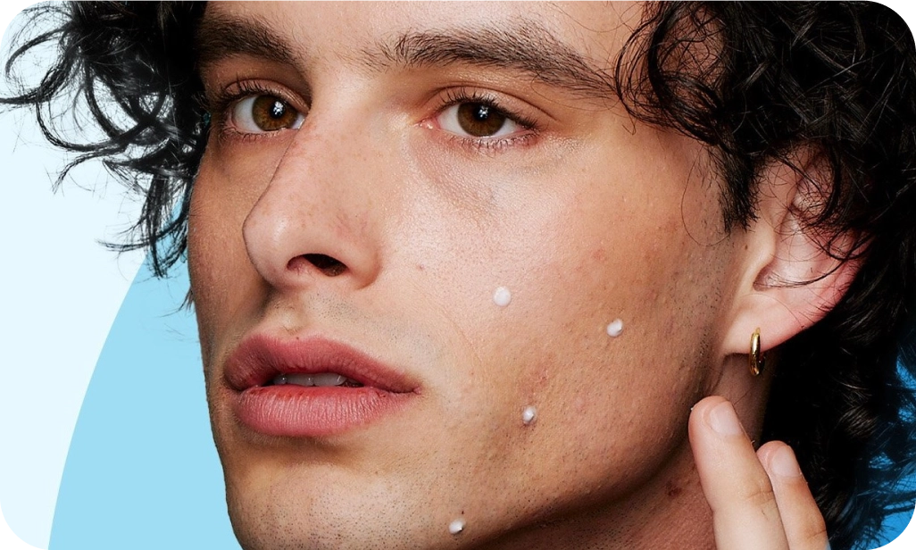 A person with curly hair applies small white dots of cream on their face, focusing intently. The background features light blue and darker blue curves.