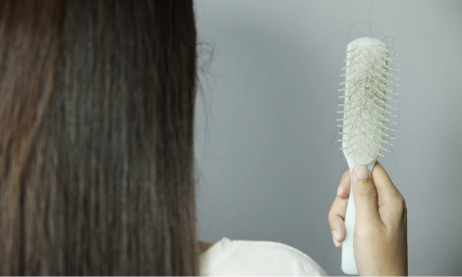 The back of a head of a woman with long brown hair as she gazes at her hair brush containing hair that has fallen out