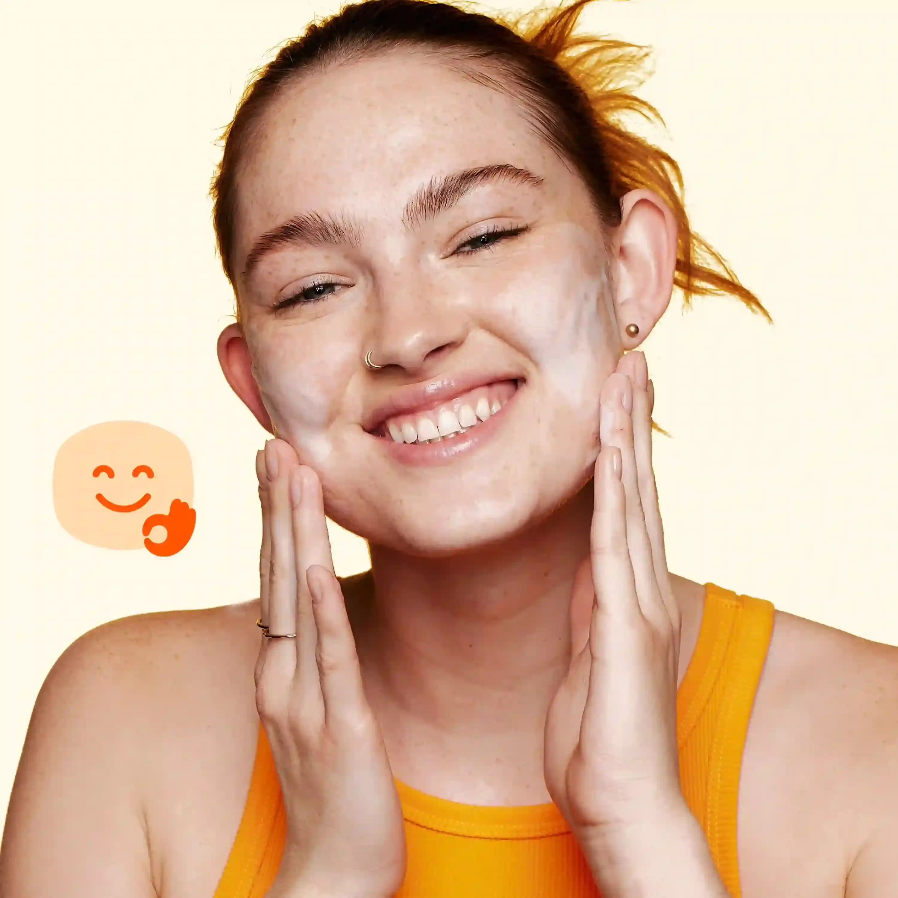 A person with their hair tied back applies facial cream while smiling, in front of a light background. A smiley face emoji with an 'okay' hand gesture is visible.