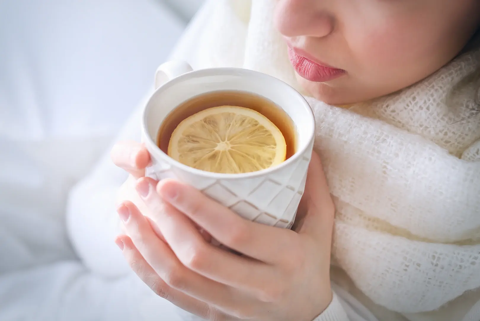 Person holding a white mug of hot tea with a lemon slice, wrapped in a white scarf, looking cozy while appearing to have a cold.