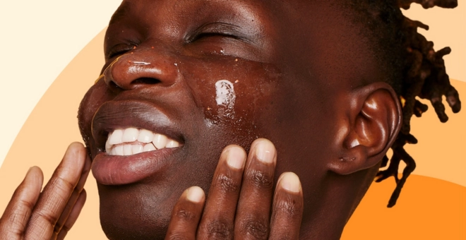 A person smiles with eyes closed, applying a shiny gel to their face. The background features soft, blended orange tones.