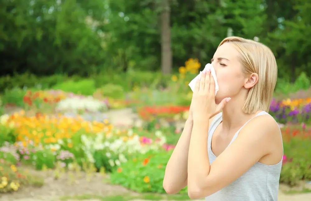 Person with short blonde hair using a tissue for a runny nose in a vibrant garden full of colorful flowers and green trees.