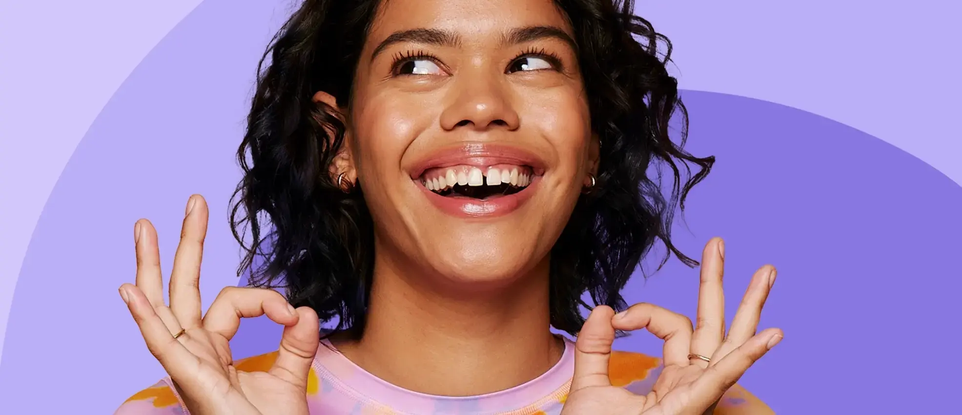 A person with curly hair smiles joyfully, making "OK" hand gestures. The background features gradient shades of purple.